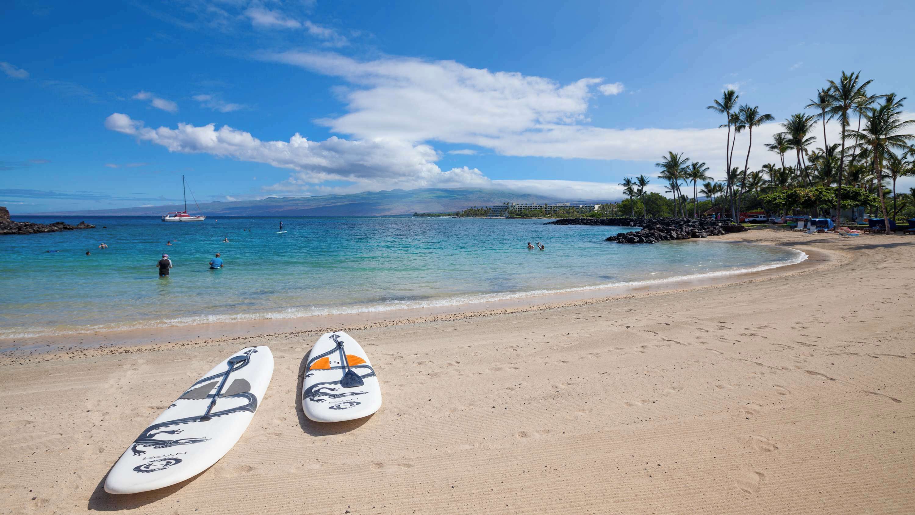 A beach with two surfboards on the sand, a clear blue sky, and a distant view of a boat and mountains.