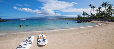A beach with two surfboards on the sand, a clear blue sky, and a distant view of a boat and mountains.