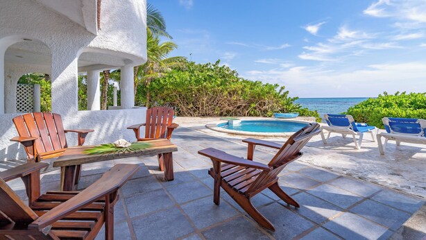 A poolside area with wooden chairs and a table, surrounded by lush greenery and a view of the ocean. - Stunning 5BR Balam Ek Villa – Oceanfront, Pool & Exceptional Views (Akumal)