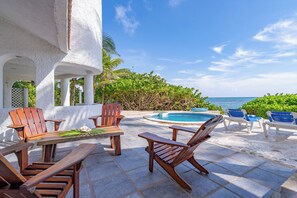 A poolside area with wooden chairs and a table, surrounded by lush greenery and a view of the ocean. - Stunning 5BR Balam Ek Villa – Oceanfront, Pool & Exceptional Views (Akumal)