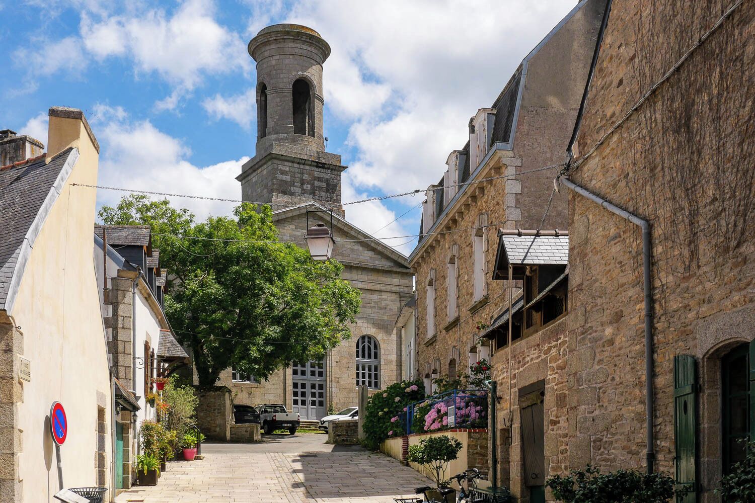 Une rue étroite bordée d’immeubles en pierre, avec une tour imposante et un arbre au centre.