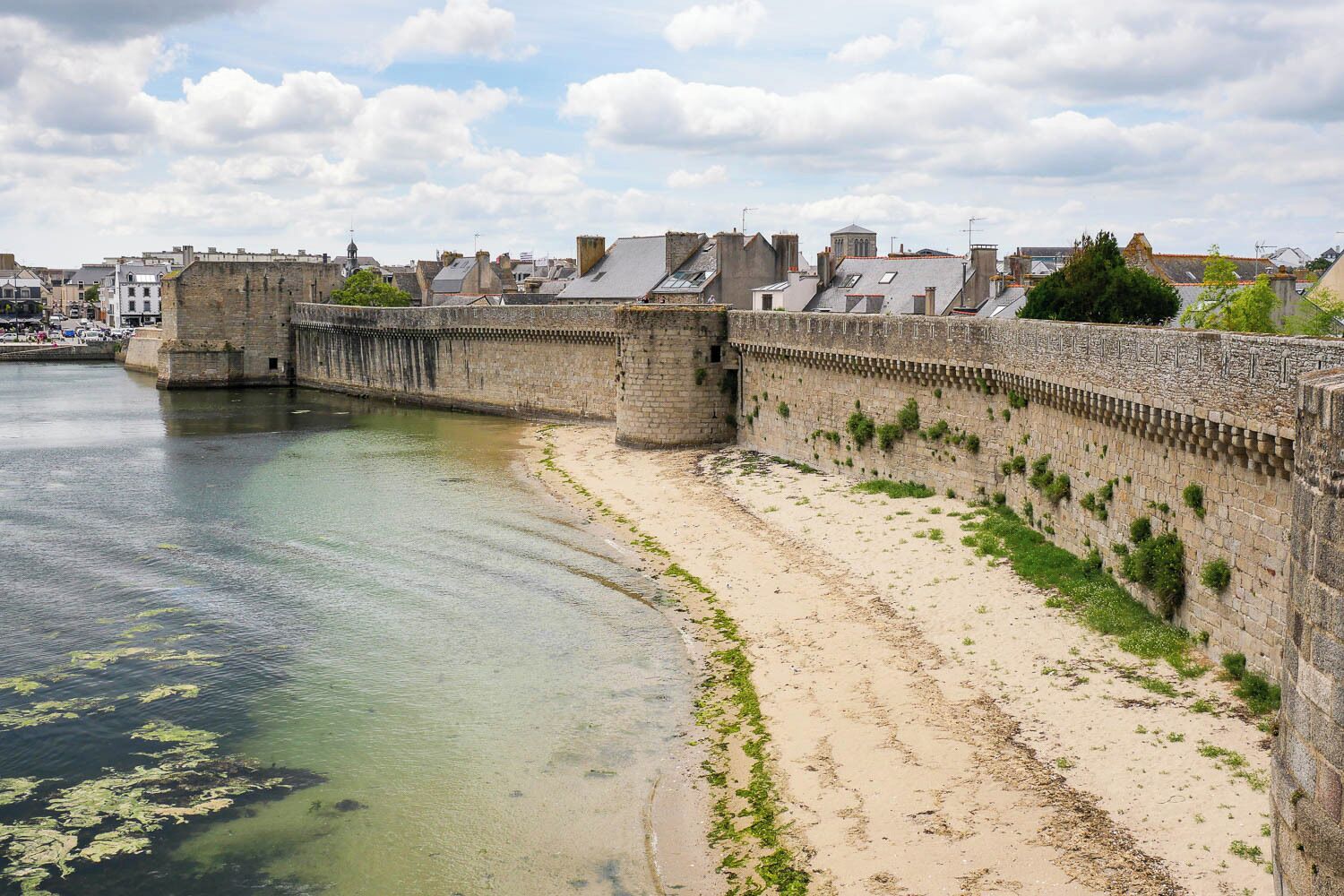 A coastal cityscape with a historic stone wall along the waterfront, clear water, and buildings in the background.