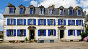 A multi-story building with a symmetrical facade, featuring a central entrance with a pediment, multiple windows with blue shutters, and a flat roof with dormer windows. - Apartment with terrace and private parking in Concarneau, Brittany (Concarneau)