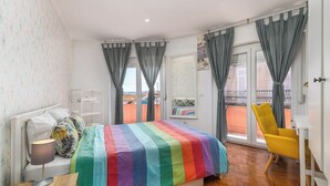 Ein Schlafzimmer mit einem bettenausgestellten Regenbogendecke, einem weißen Nachttisch mit Lampe, einem gelben Sessel und einem Balkon mit grauen Vorhängen.