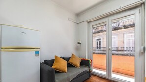 Ein kleines Wohnzimmer mit einer Couch, einem Kühlschrank und Blick auf einen Balkon mit Geländer.