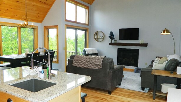 A spacious living area with a kitchen island, wooden flooring, a fireplace, and large windows offering a view of the outdoors. - Rock Pond Condo 53 - Ski in Ski out Rock Pond Condo at Saddleback (Rangeley)