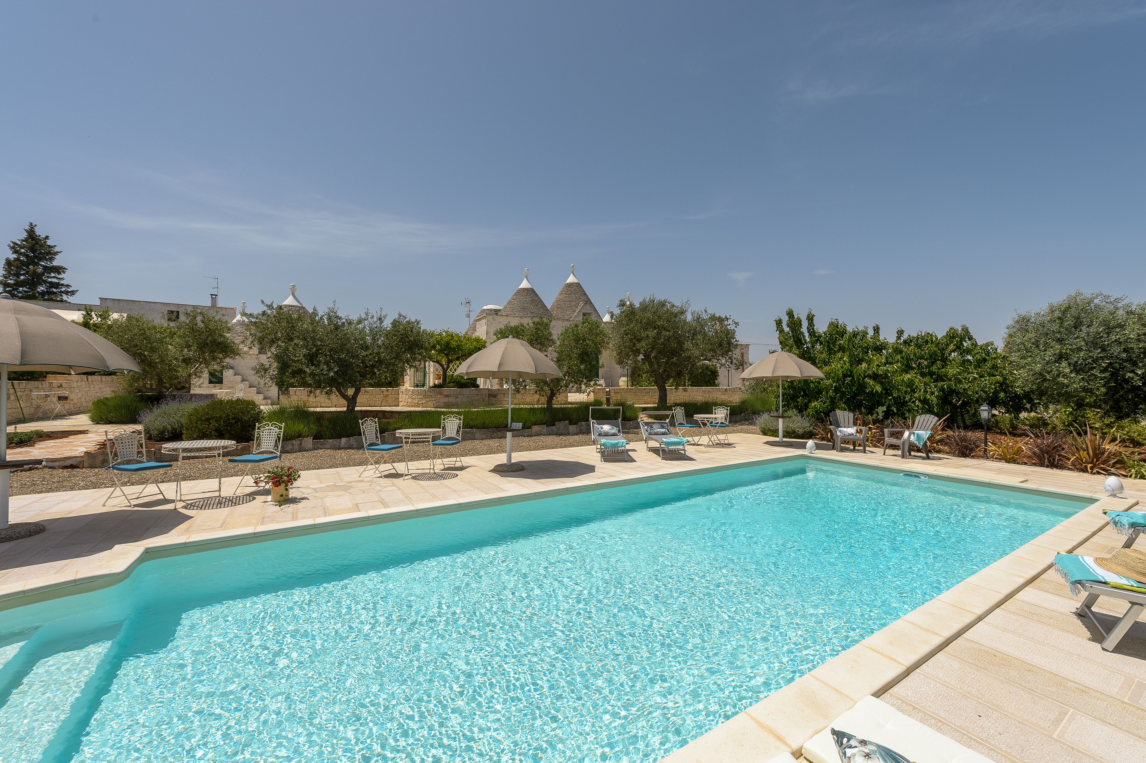 A swimming pool with lounge chairs and umbrellas, surrounded by greenery and buildings with domed roofs.