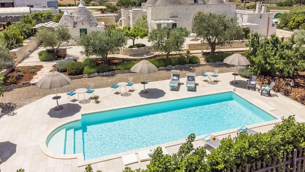 Piscina con sillas de descanso y sombrillas, rodeada de edificios de piedra y vegetación.