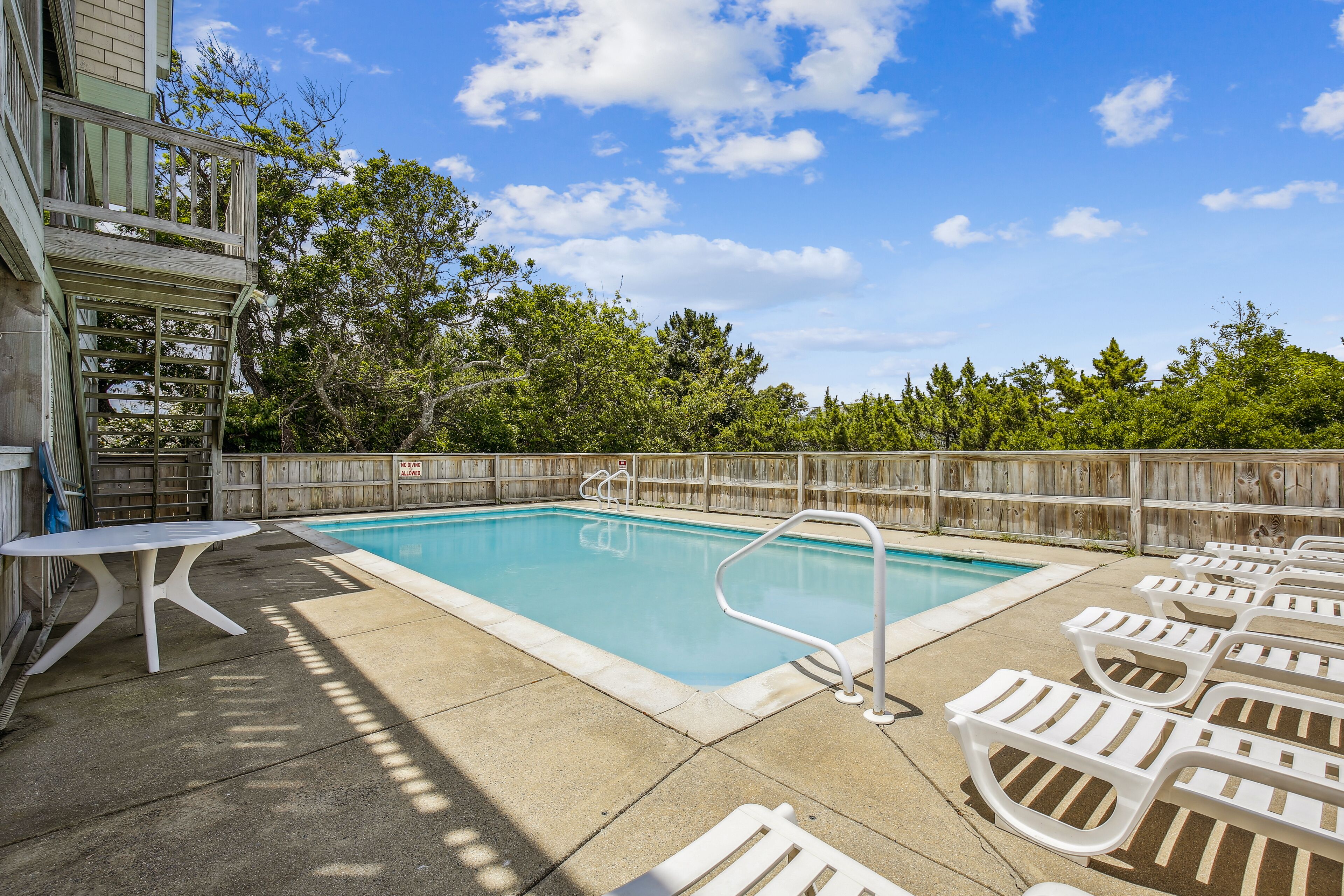 A swimming pool with lounge chairs and a table, surrounded by a wooden fence and trees.