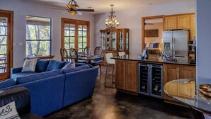 Een ruime woonkamer met een grote blauwe bank, een eethoek met houten stoelen, een keuken met een koelkast en kastjes, en een kroonluchter.