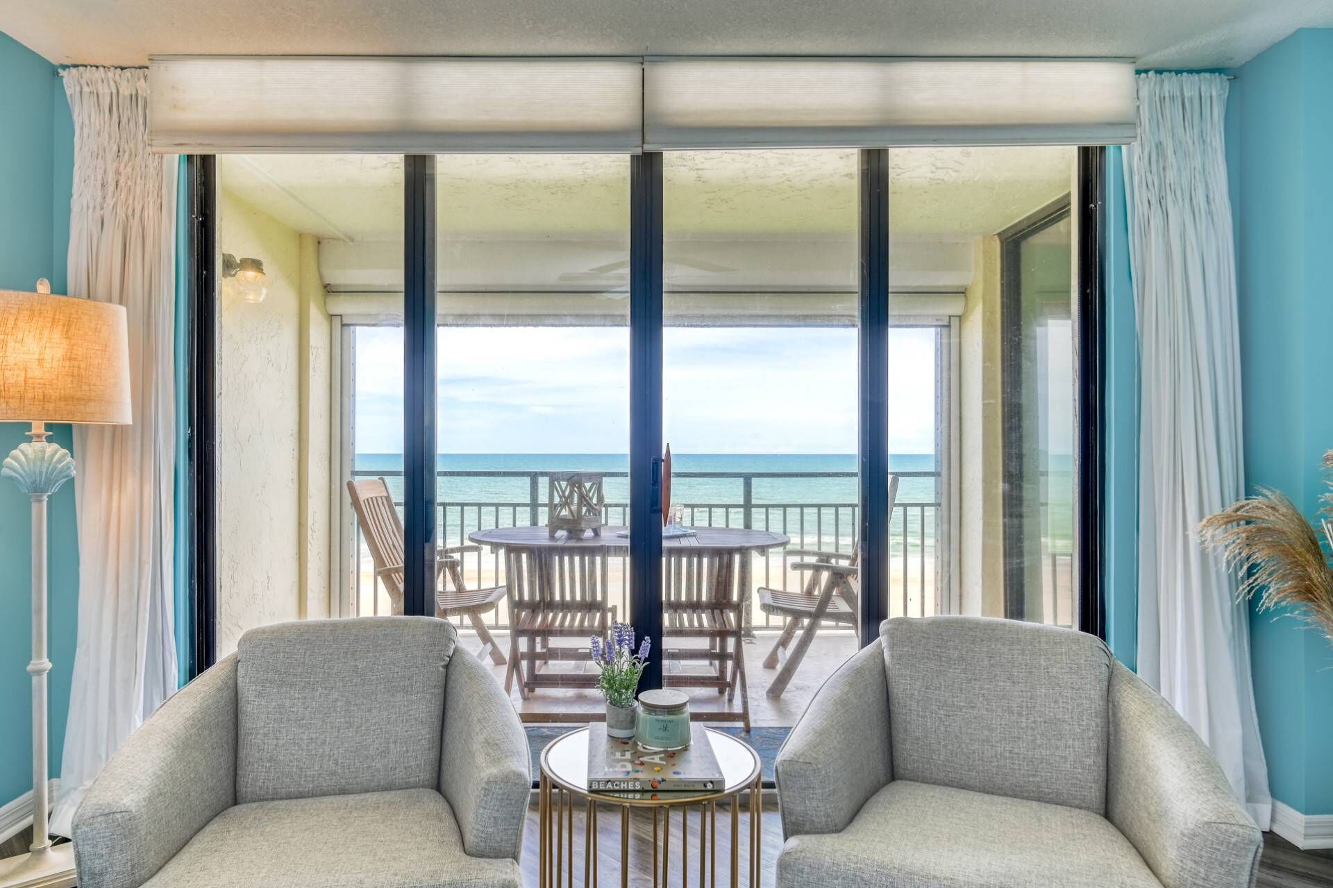 A room with two grey sofas, a small round table with a lamp, and a view of the ocean through large windows.