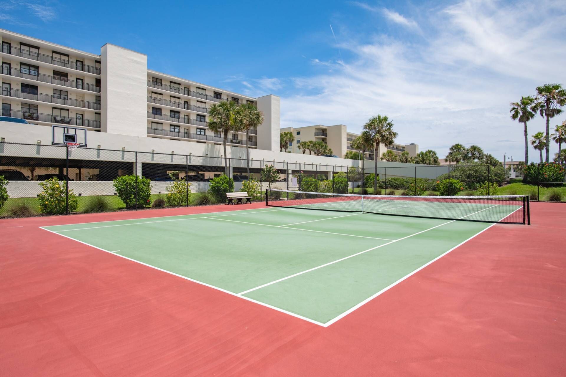 Een buitenbaan voor tennis met een net, omgeven door een hek en palmbomen.