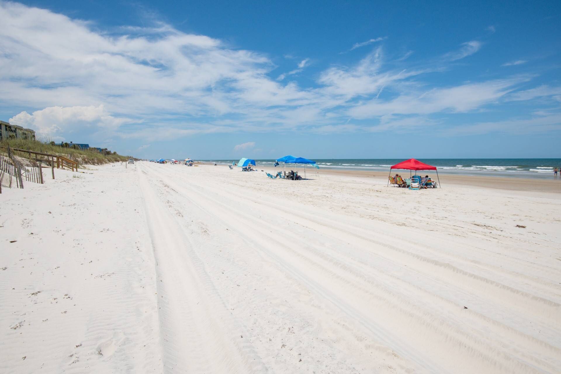 Une plage avec un sentier de sable, des parasols bleus et rouges, et une clôture en bois.