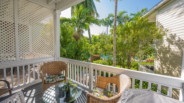 Une véranda couverte, meublée de meubles en osier et dotée d’une table en verre, offre une vue sur un jardin luxuriant, avec des palmiers et une maison en arrière-plan.
