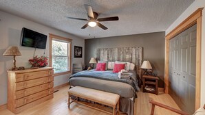 A bedroom with a bed, wooden dresser, bench, ceiling fan, and a TV on the wall.