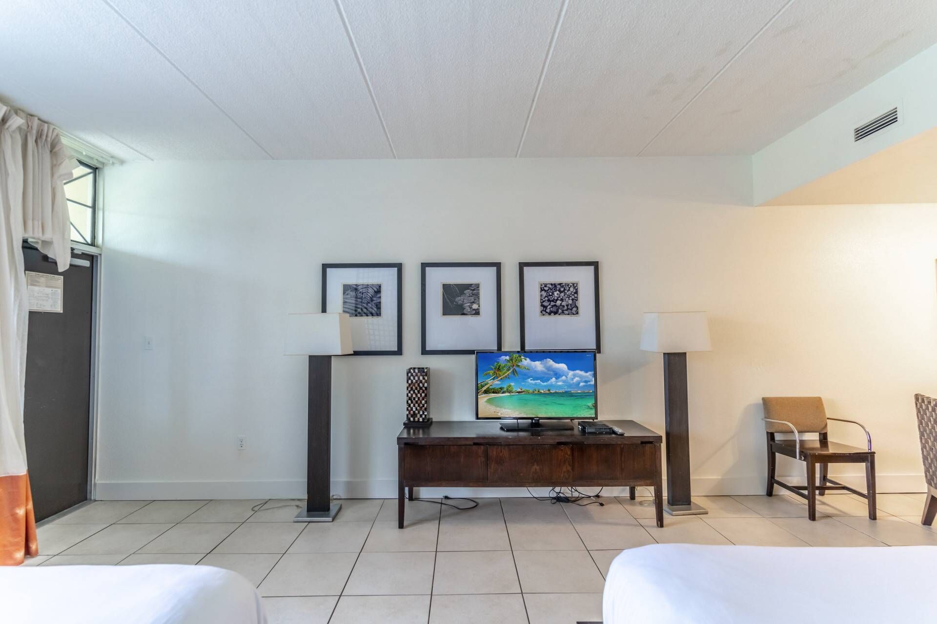 A hotel room with a bed, a desk with a computer, a chair, and three framed pictures on the wall.