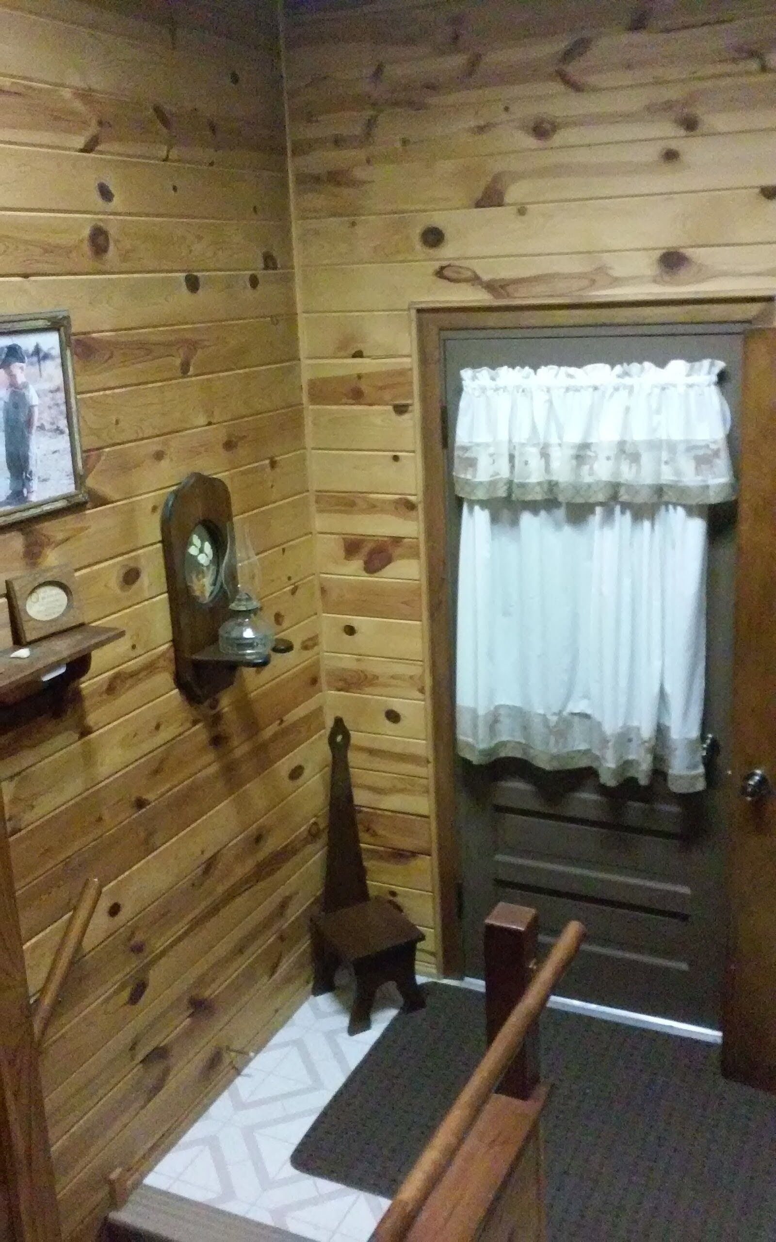 A wooden cabin interior with a wooden door, a small stool, a mirror, and a framed picture on the wall.