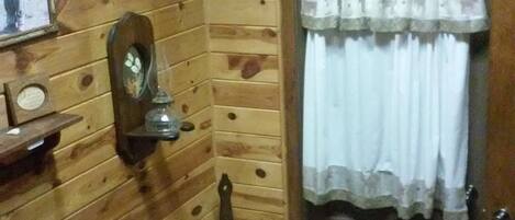 A wooden cabin interior with a wooden door, a small stool, a mirror, and a framed picture on the wall.