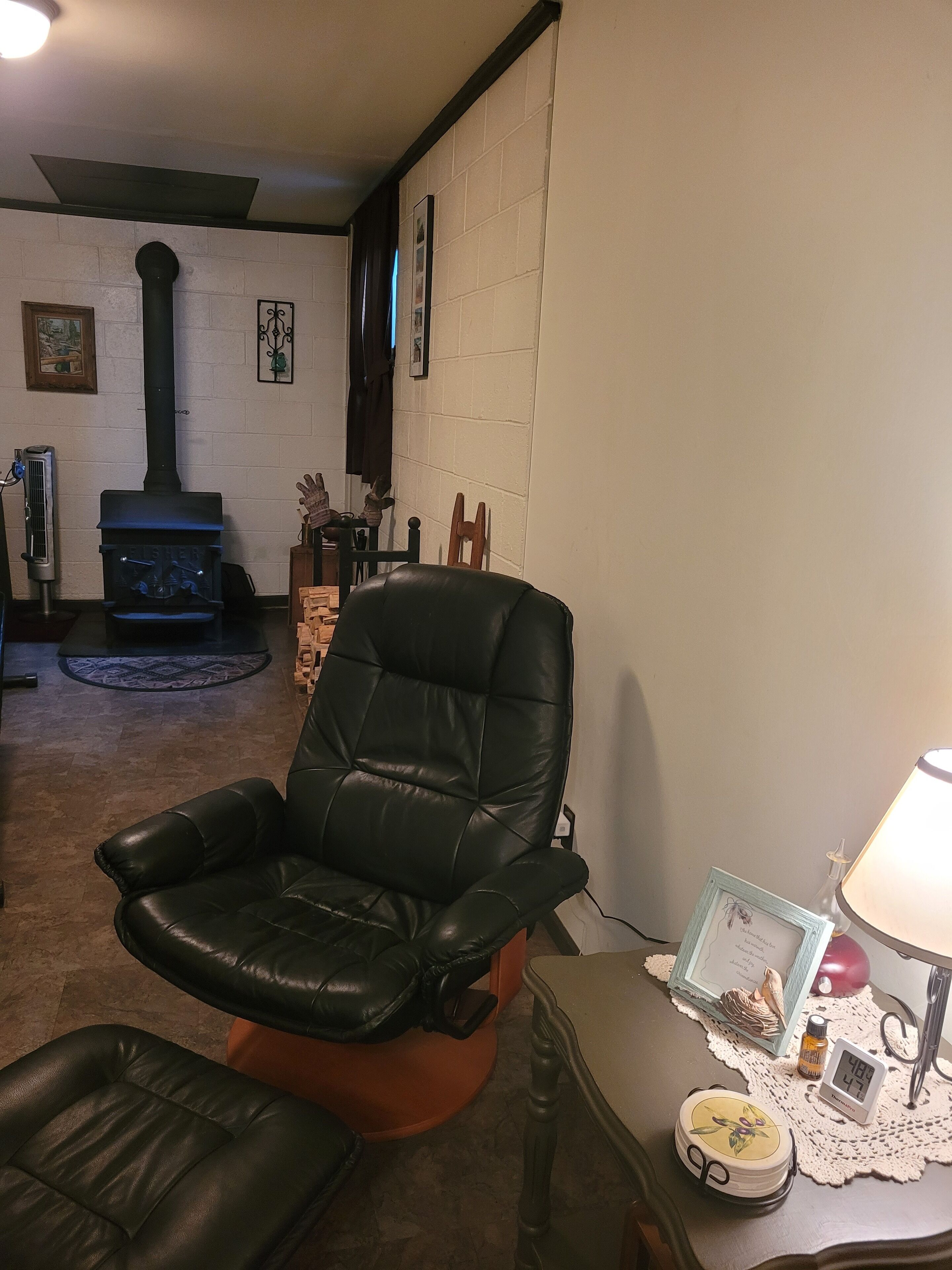A living room with a black leather chair, a small table with a clock and decorative items, a fireplace, and a fan.