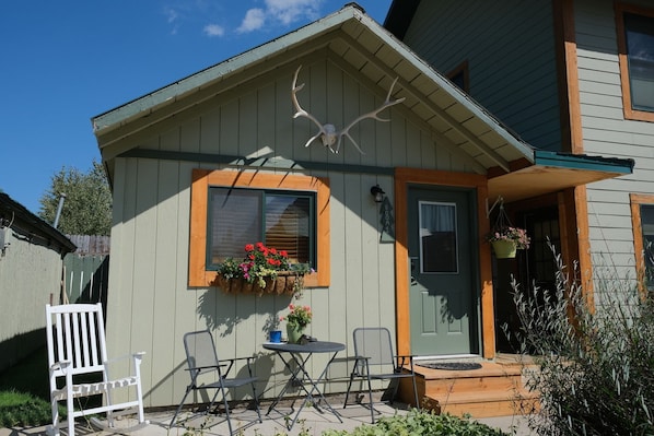 A small house with a porch, a wooden rocking chair, a table, and a flower box with red flowers.