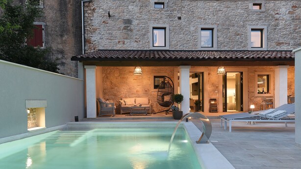 Una piscina con un edificio de piedra al fondo, un área de patio con sillones de descanso y una pared de piedra.