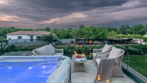 Terraza en la azotea con jacuzzi, mobiliario de mimbre y balaustrada de vidrio, con vistas a un jardín exuberante y una casa.