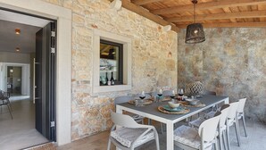 Comedor con paredes de piedra, una mesa preparada para cuatro personas, sillas blancas y una lámpara colgante.