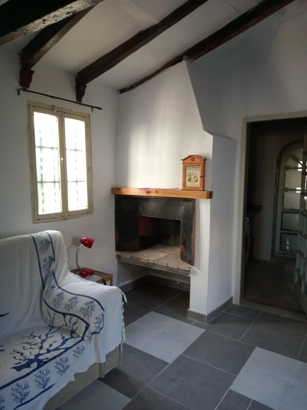 A room with a tiled floor, a bench with a patterned cover, a red lamp, a fireplace with a wooden mantel, and a door leading to another room.