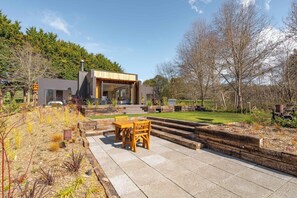 Una casa moderna con un patio attrezzato con tavolo e sedie in legno, immersa in un giardino con diverse piante e un prato curato.