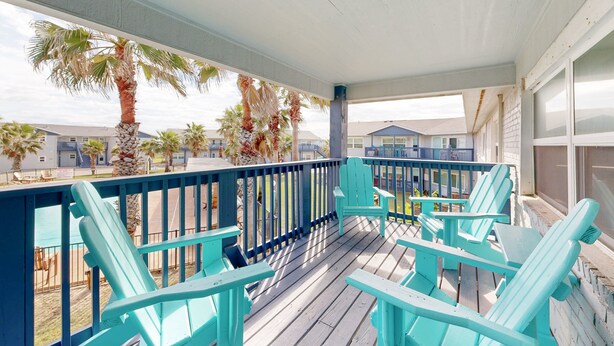 Une véranda couverte avec des fauteuils Adirondack turquoise, une terrasse en bois et une vue sur les palmiers et les maisons au loin.