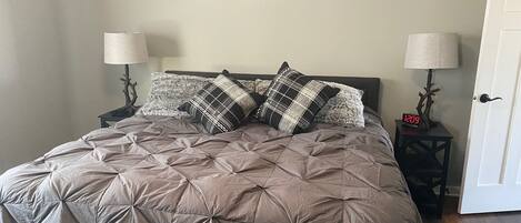 Una cama bien tendida con una colcha marrón y dos almohadones decorativos, flanqueada por dos mesitas de noche con lámparas.