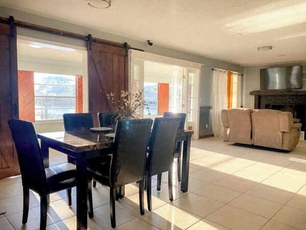 A dining area with a table and chairs, a fireplace, and a sofa in a well-lit room with large windows.