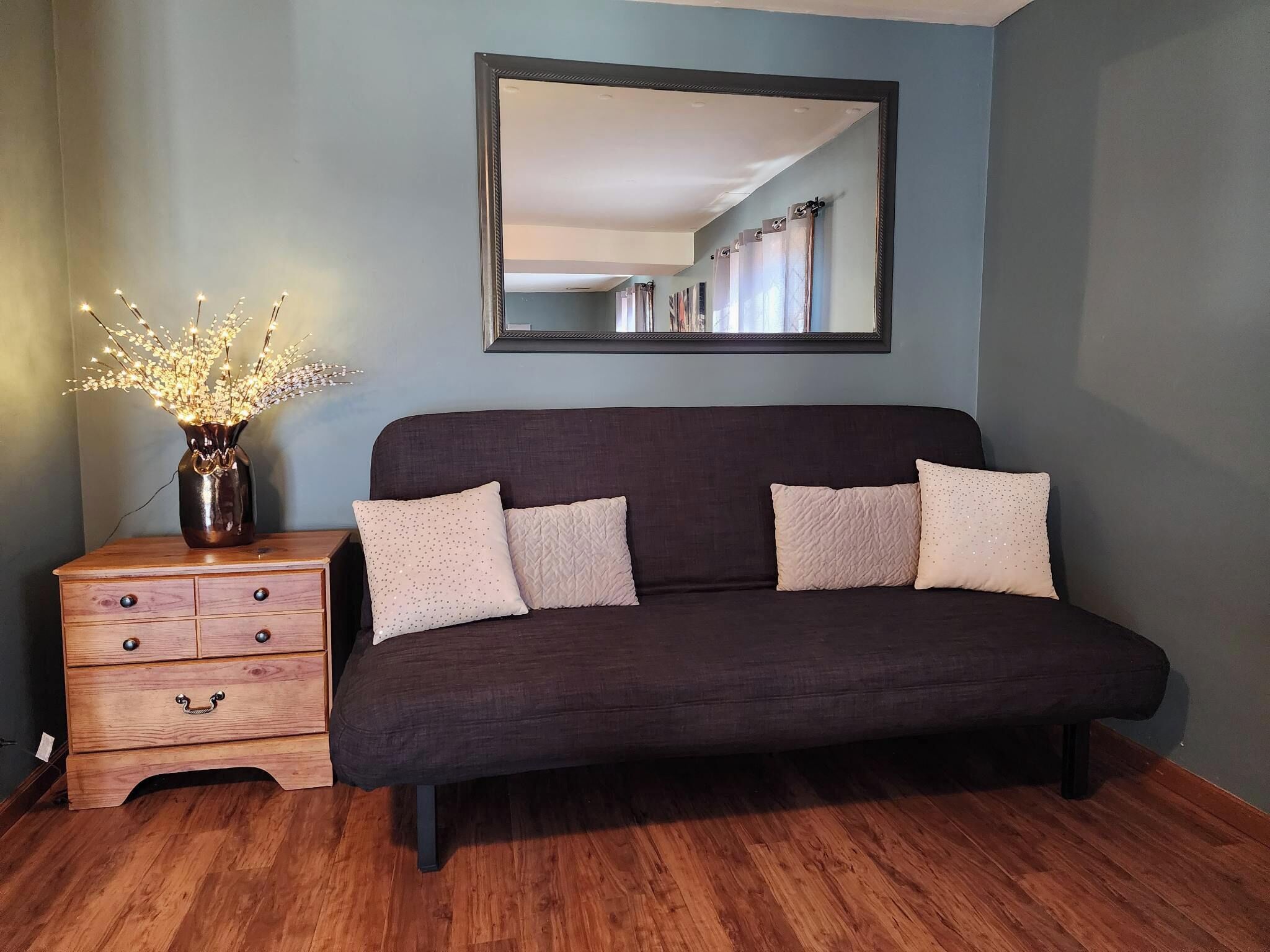 Ein Zimmer mit einem dunkelgrauen Sofa, einer Holzkommode mit Schubladen, einer Vase mit Beleuchtung und einem Spiegel an der Wand.