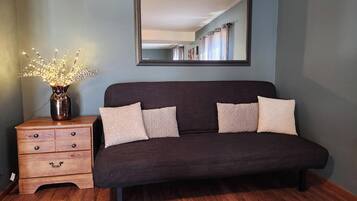 Ein Zimmer mit einem dunkelgrauen Sofa, einer Holzkommode mit Schubladen, einer Vase mit Beleuchtung und einem Spiegel an der Wand.