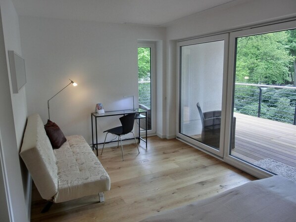 A modern bedroom with a bed, a desk, and a chair. There is a large window providing a view of greenery outside.