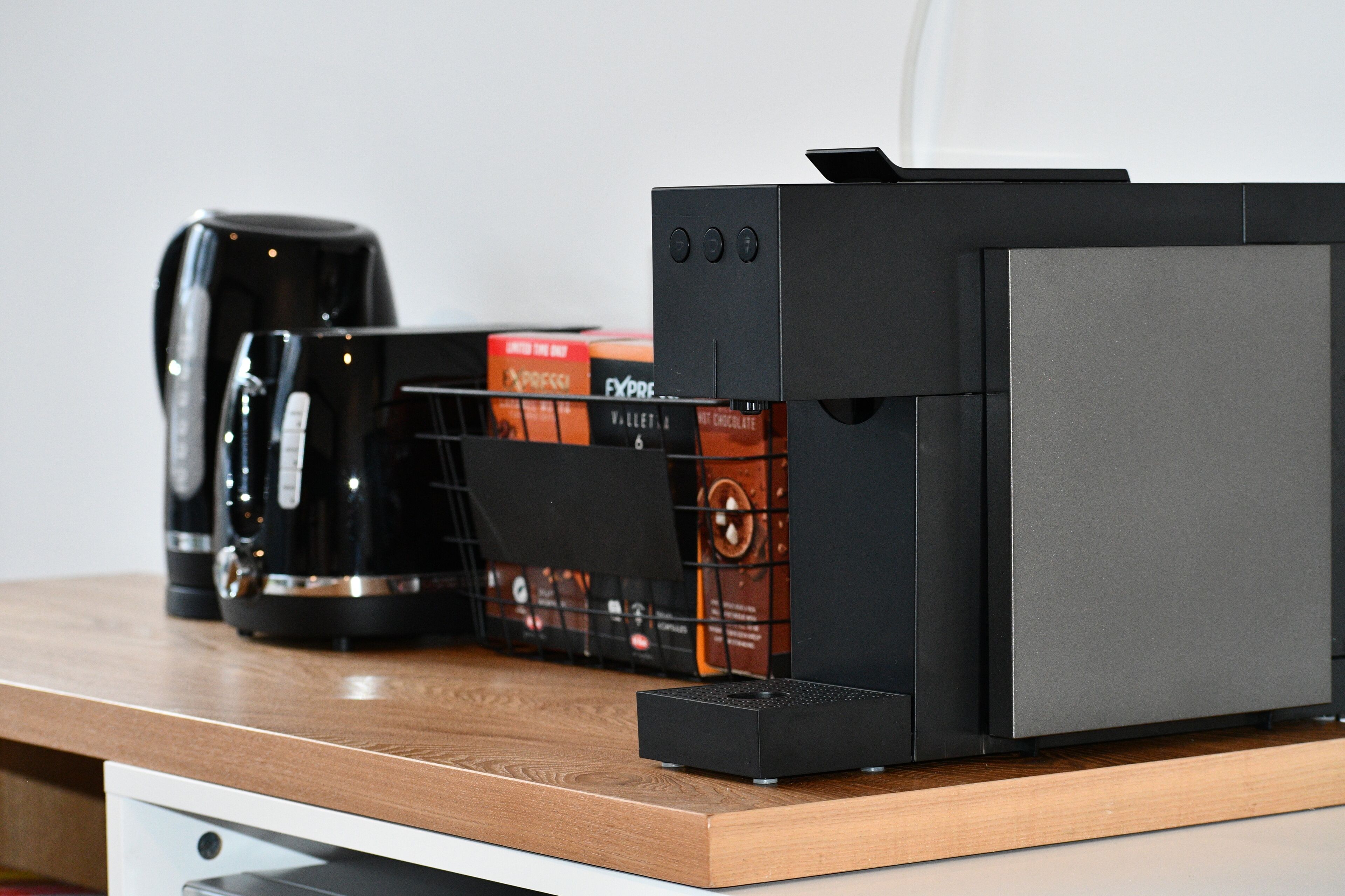 A coffee machine, a box of coffee pods, and a black electronic device on a wooden surface.