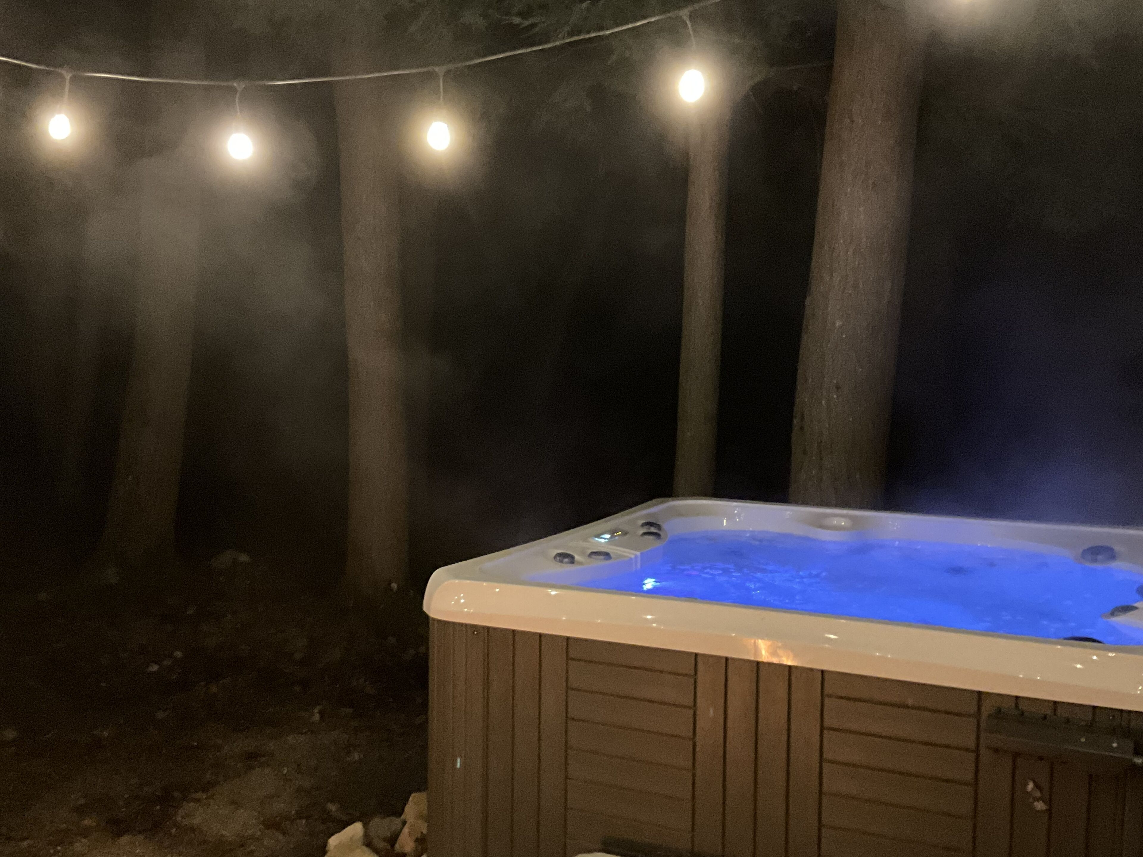 A hot tub with a wooden exterior, surrounded by trees and string lights.