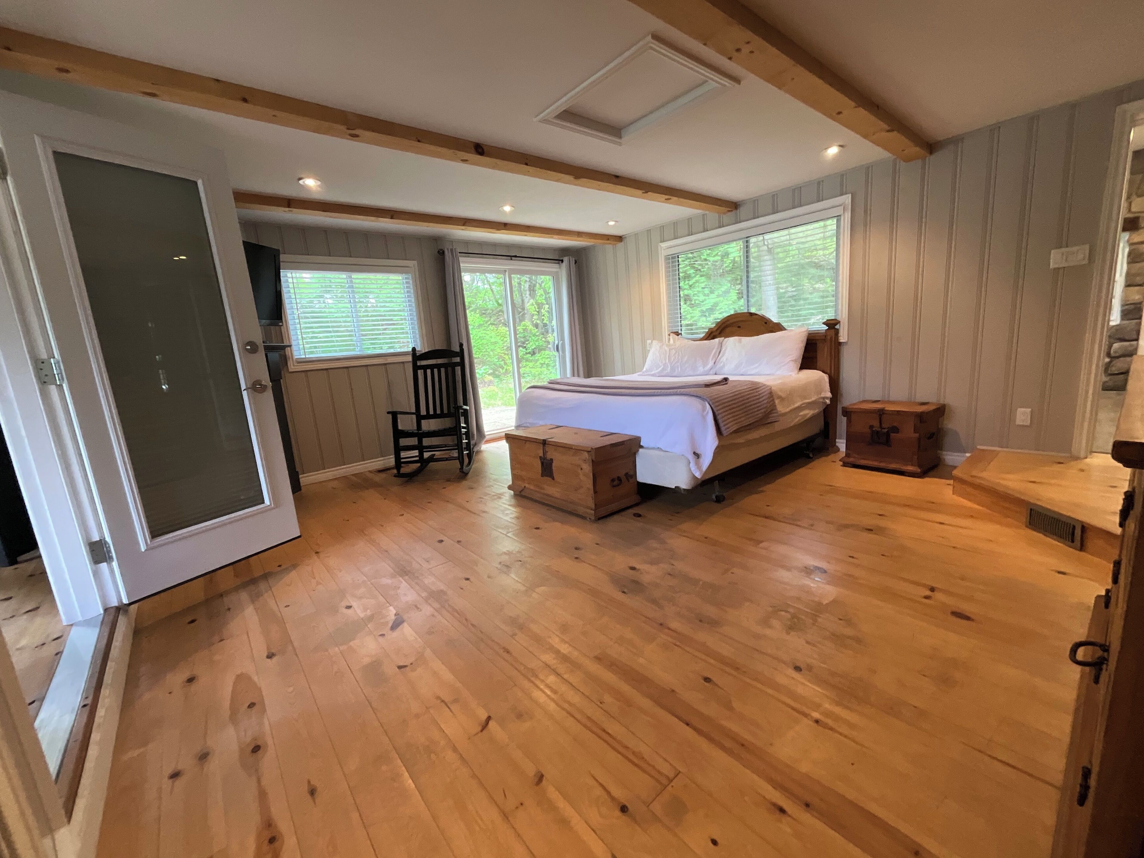 A bedroom with a wooden floor, a bed with white bedding, a rocking chair, and a window with blinds.