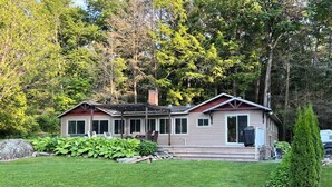 A two-story cabin with a covered patio, surrounded by a well-maintained lawn and a small pond. - Bright and spacious lakeside cottage with firepit, hot tub and AC on Stoney Lake (Lakefield)