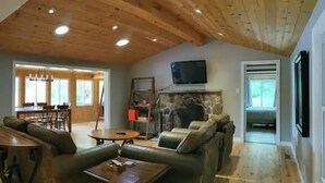 A spacious living room with a high wooden ceiling, a stone fireplace, leather sofas, and a dining area in the background. - Bright and spacious lakeside cottage with firepit, hot tub and AC on Stoney Lake (Lakefield)
