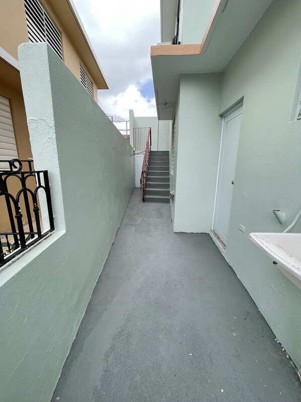 Une ruelle étroite avec un sol en béton, une rambarde en métal et un escalier menant à une porte.
