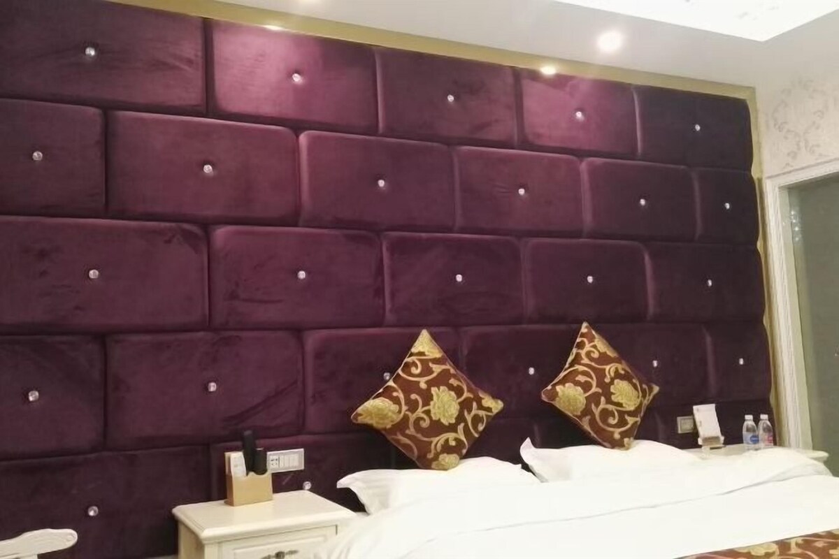 A hotel room with a large, tufted headboard, a neatly made bed, a bedside table with a lamp, and a small nightstand.