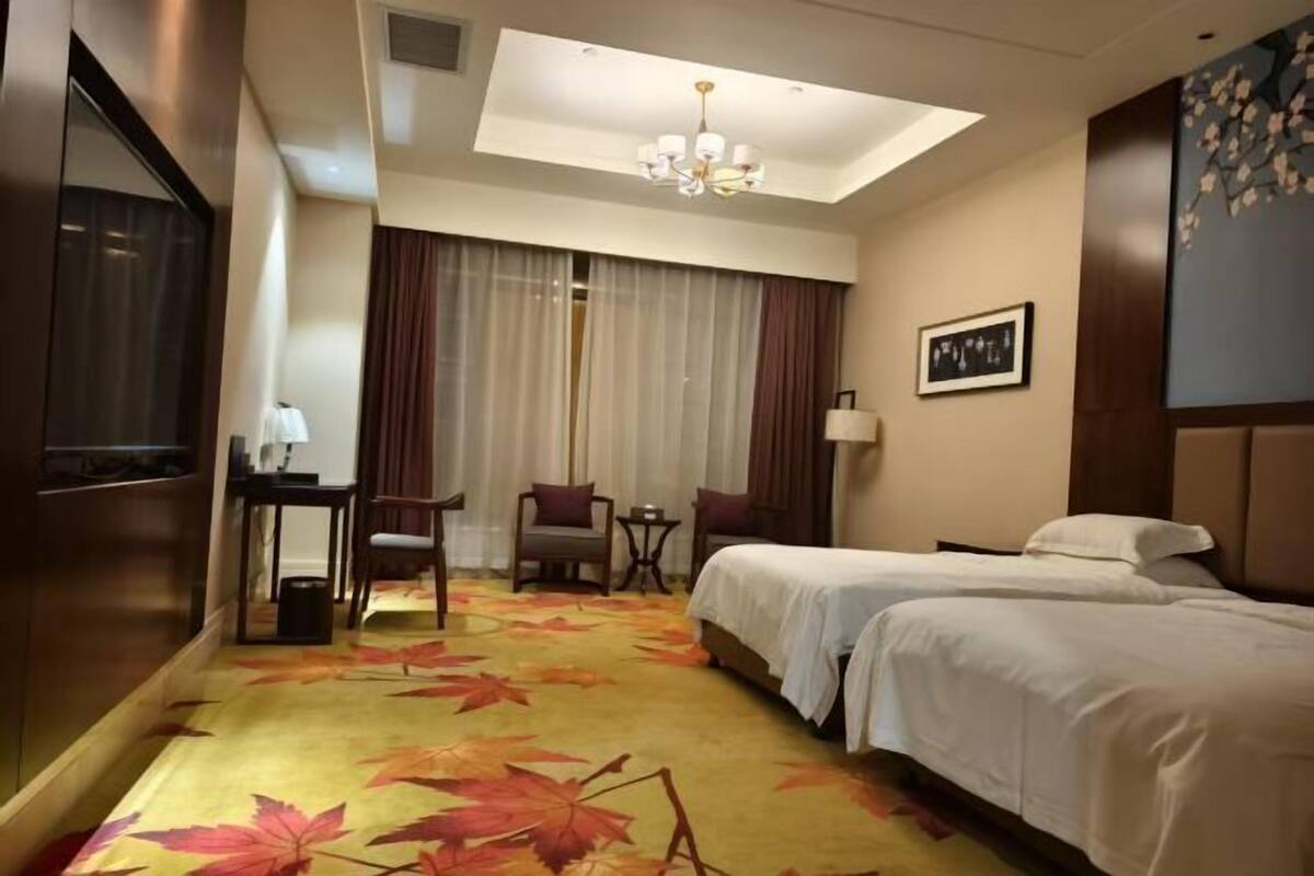 A hotel room with two beds, a desk, a chair, a small table, a chandelier, and a large window with curtains.