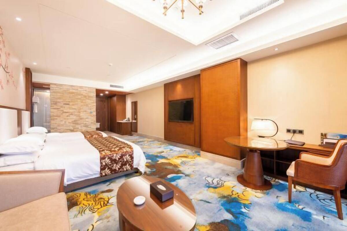 A hotel room with a bed, a desk with a lamp, a chair, a small table, and a TV.