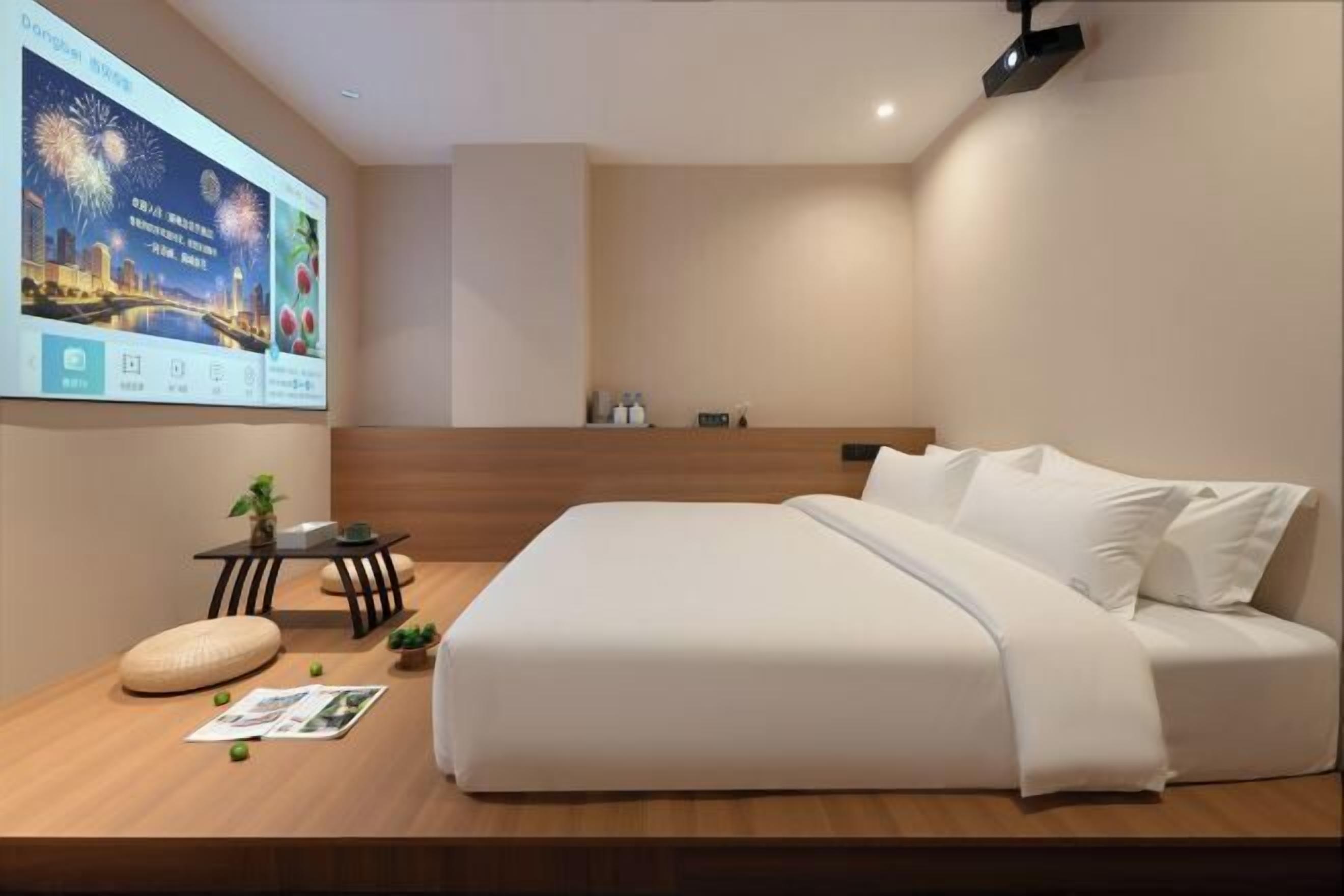 A modern hotel room with a large bed, a small table with a plant, a wall-mounted TV, and a wooden platform with a magazine and a pillow.