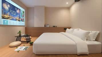 A modern hotel room with a large bed, a small table with a plant, a wall-mounted TV, and a wooden platform with a magazine and a pillow.