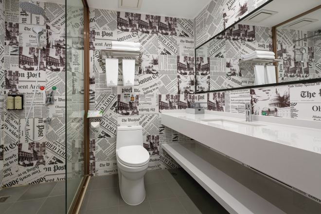 Baño con inodoro blanco, espejo y lavamanos. Las paredes están cubiertas con recortes de periódicos.