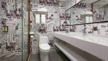 Baño con inodoro blanco, espejo y lavamanos. Las paredes están cubiertas con recortes de periódicos.