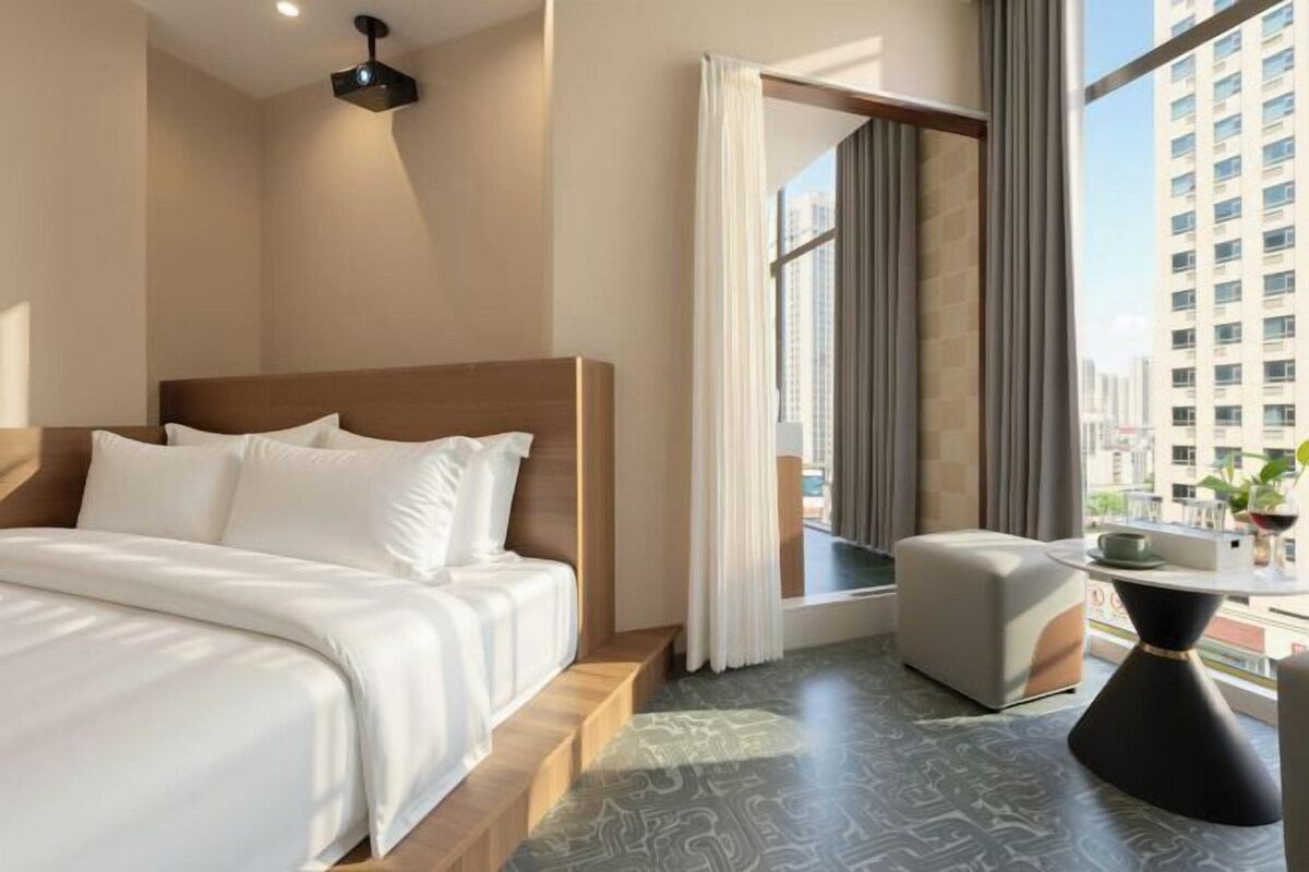 Habitació d'hotel amb llit, taula petita, cadira, finestra amb cortines i vista a un paisatge urbà.