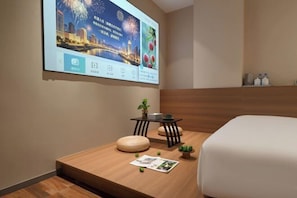 Habitación de hotel con cama, una mesita con una planta, una pantalla que muestra un paisaje urbano y un banco de madera.